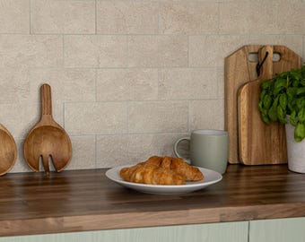 May include: A kitchen scene featuring a dark wood countertop with a white plate holding two croissants, a light green mug, wooden salad servers, two wooden cutting boards, and a potted green plant against a beige tiled backsplash.