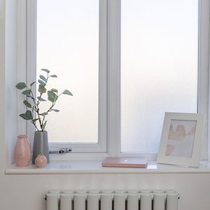 May include: A white window frame with frosted glass panes sits above a white radiator. On the windowsill are two pink vases, one grey textured vase with a green leafy branch, a small pink notebook, and a white picture frame with abstract art.