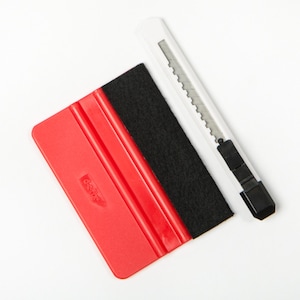 May include: A red plastic squeegee with a black felt edge and a white retractable utility knife with a silver metal blade. The squeegee features two parallel vertical ridges and a small embossed logo on its surface, positioned next to the knife.