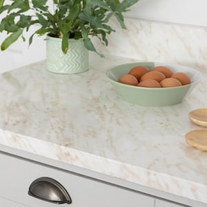 Puede incluir: Una encimera de cocina con un patrón similar al mármol en tonos blancos y beige. Un cuenco verde claro lleno de huevos marrones se encuentra sobre la encimera, junto con una pequeña planta verde en una maceta decorativa. También se ven cucharas de madera.