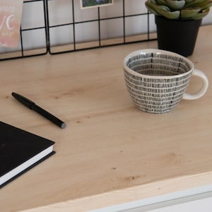 May include: A black and white patterned ceramic coffee mug with a white handle sits on a light wood surface. A black pen and a black notebook are also on the surface. A small potted plant and a wire grid are in the background.