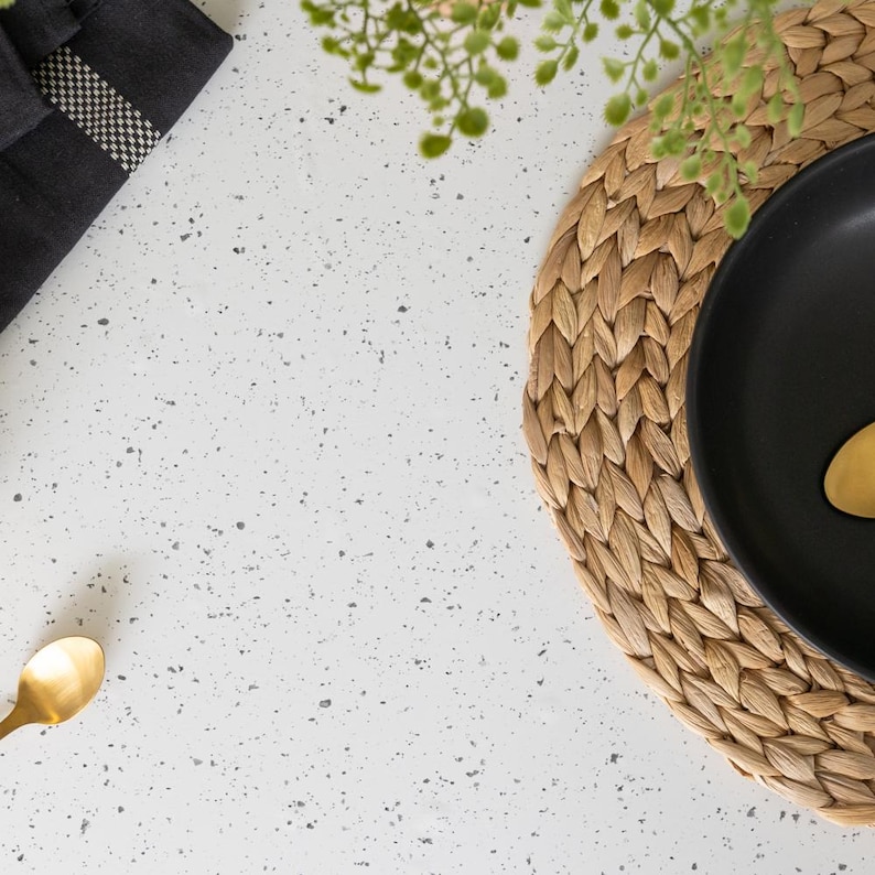 May include: Overhead shot of a table setting with a black plate, gold spoon, and woven placemat. A dark gray napkin with a checkered pattern is in the upper left corner. Green sprigs add a natural touch to the composition.