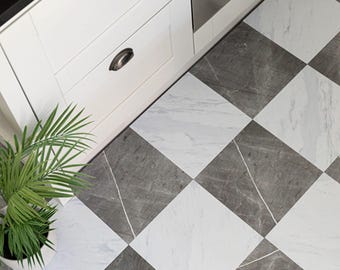May include: Square floor tiles arranged in a checkerboard pattern featuring alternating white and dark gray marble textures with visible veining. The tiles are installed on a kitchen floor next to white cabinets with dark metal drawer pulls.