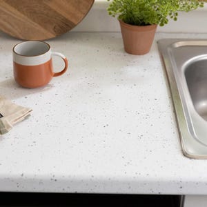 May include: A kitchen countertop with a speckled white surface. A brown and white mug sits on the counter, next to a small potted plant and a stainless steel sink. A wooden cutting board is in the background.