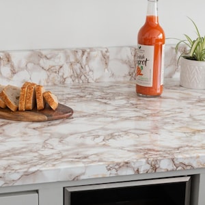 May include: A kitchen countertop with a marble effect. A bottle of carrot juice and a wooden board with sliced bread are on the surface. A small potted plant is also visible.