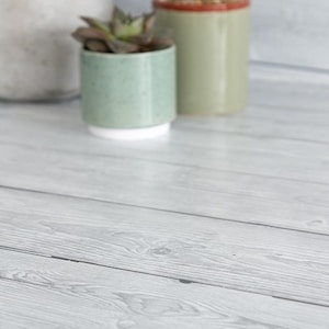 Puede incluir: Una superficie de madera gris claro con vetas visibles. En el fondo, hay tres pequeñas macetas de cerámica. Una es verde claro con base blanca, otra es verde claro y la tercera es beige claro con borde marrón. Las macetas contienen pequeñas plantas verdes.