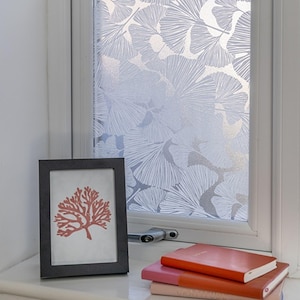 May include: A window covered in a translucent film featuring a white ginkgo leaf pattern. In front of the window sits a black picture frame containing a coral-coloured coral illustration and a stack of three notebooks in orange, pink, and peach.