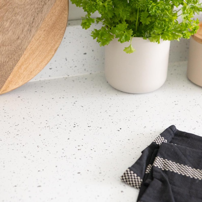 May include: A kitchen countertop scene featuring a wooden cutting board, a white ceramic pot with green herbs, and a black and white striped kitchen towel. The countertop is white with speckled details.