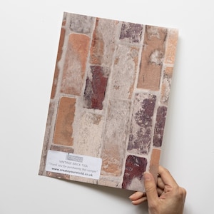 May include: A sample of vintage brick wallpaper with a distressed, weathered look. The wallpaper features a mix of red, brown, and beige bricks with white mortar. The sample is labelled "CREATE YOUR WORLD" and "Vintage Brick Tisa".