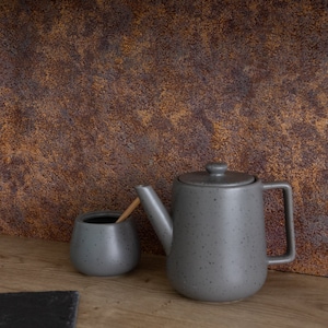Op de afbeelding: Een grijze keramische theepot en suikerpot met een houten roerder op een houten oppervlak voor een roestige bruine achtergrond.