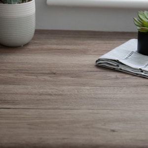 Puede incluir: Primer plano de una superficie de madera de color marrón claro con vetas. Un paño a rayas blancas y grises está doblado sobre la superficie, con una pequeña maceta negra que contiene una planta verde. También se ve una maceta de cerámica blanca con una planta.