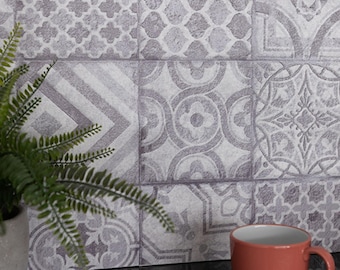 May include: A wall covered in square tiles featuring a variety of gray and white geometric and floral patterns. A green fern in a gray pot sits to the left, and a solid orange mug is positioned on a dark surface in the lower right corner.
