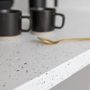 May include: A close-up of a white countertop with a speckled pattern. Black mugs with beige bottoms are in the background, along with a gold spoon. The scene suggests a kitchen or dining setting.