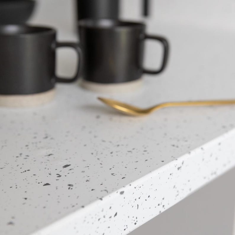 May include: A close-up of a white countertop with a speckled pattern. Black mugs with beige bottoms are in the background, along with a gold spoon. The scene suggests a kitchen or dining setting.