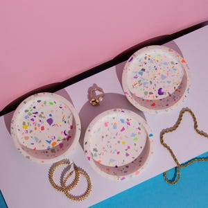 Könnte beinhalten: Drei runde weiße Schalen mit einem mehrfarbigen Terrazzo-Muster aus rosa, blauen, orangefarbenen, grünen und violetten Sprenkeln. Das Set wird durch einen goldfarbenen Ring mit Blumenmotiv, ein Paar goldene Creolen und eine goldene Kette ergänzt.