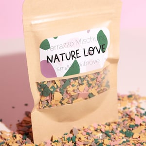Könnte beinhalten: Ein Kraftpapierbeutel mit einem transparenten Fenster, gefüllt mit bunten Terrazzo-Chips. Das Etikett lautet "Terrazzo Mischung NATURE LOVE smartwithove". Die Chips sind eine Mischung aus Grün, Gelb, Rosa und Beige.