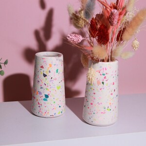 Könnte beinhalten: Zwei zylindrische weiße Vasen mit einem mehrfarbigen Terrazzo-Muster aus neonpinken, orangen, gelben, blauen und grünen Sprenkeln. Eine Vase ist leer, während die andere mit einem Strauß getrockneter Gräser in Rosa-, Rot- und Beigetönen gefüllt ist.