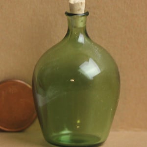 Könnte beinhalten: Kleine, olivgrüne Glasflasche mit Korken. Die Flasche hat einen abgerundeten Körper und einen schmalen Hals. Der Korken ist hellbraun. Die Flasche ist leer und scheint aus der Zeit zu stammen.