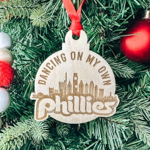 Puede incluir: Un adorno navideño de madera con una cinta roja. El adorno tiene forma de círculo y tiene un horizonte de Filadelfia, Pensilvania con el texto "Dancing on my own Phillies" alrededor del horizonte.