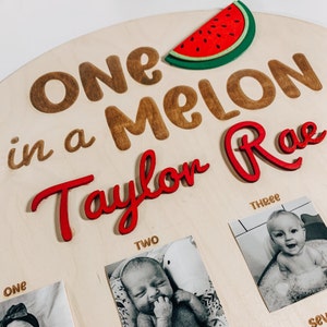 Może przedstawiać: Okrągły drewniany znak z napisem "One in a Melon Taylor Rae" w brązowych i czerwonych literach. Nad tekstem znajduje się czerwony plaster arbuza. Pod tekstem znajdują się cztery ramki na zdjęcia z numerami "One", "Two", "Three" i "Seven". Każda ramka zawiera czarno-białe zdjęcie dziecka.