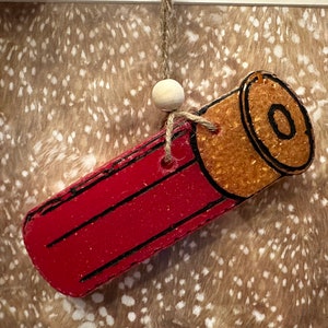 May include: A red and gold shotgun shell ornament featuring black outline details. The ornament is suspended by a piece of twine with a small, round, light-colored wooden bead attached above the shell for decorative hanging.
