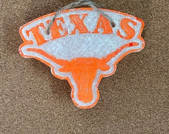 May include: An orange and white Texas Longhorns ornament with the word "TEXAS" in orange block letters. The ornament features the Longhorns logo and is suspended by a string with a wooden bead.
