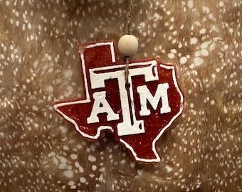 May include: A red and white Texas-shaped car air freshener with the letters "ATM" in white. The air freshener is hanging from a string with a wooden bead.