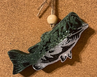 May include: A green and white bass fish ornament with a glitter finish. The ornament is hanging from a brown twine string with a wooden bead.
