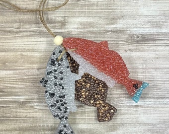 May include: Three fish-shaped car air fresheners hanging from a string. The fish are layered on top of each other. The top fish is red, the middle fish is brown, and the bottom fish is white with black spots.