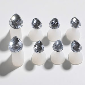 May include: Eight clear glass teardrop-shaped rhinestones on white silicone bases. The rhinestones have a faceted design and reflect light.