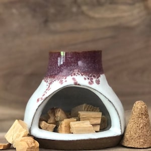 Puede incluir: Quemador de incienso de cer&aacute;mica blanca con un borde marr&oacute;n y un esmalte morado moteado. El quemador tiene una gran abertura en la parte delantera para sostener varillas o conos de incienso. El quemador est&aacute; lleno de astillas de madera y un cono de incienso marr&oacute;n est&aacute; sentado a su lado.