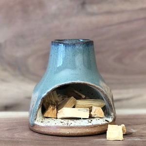 Puede incluir: Un quemador de incienso de cer&aacute;mica con un esmalte azul claro y un borde blanco. El quemador tiene una peque&ntilde;a abertura en la parte delantera para sostener varillas de incienso. El quemador est&aacute; lleno de astillas de madera.
