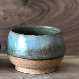 May include: A small, handmade ceramic bowl with a light blue glaze and a brown rim. The bowl has a speckled, rustic look and is perfect for serving small snacks or desserts.
