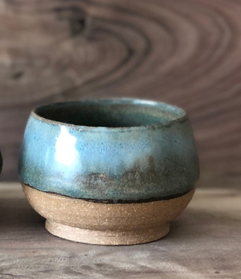 May include: A small, handmade ceramic bowl with a light blue glaze and a brown rim. The bowl has a speckled, rustic look and is perfect for serving small snacks or desserts.