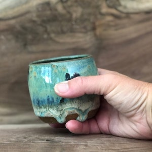 May include: A small, round, ceramic cup with a light blue glaze and brown accents. The cup has a slightly uneven rim and a drip glaze effect.