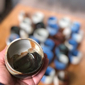 May include: A small, brown and white ceramic bowl with a dark brown glaze inside. The bowl is being held in a hand, and there are many other similar bowls in the background.