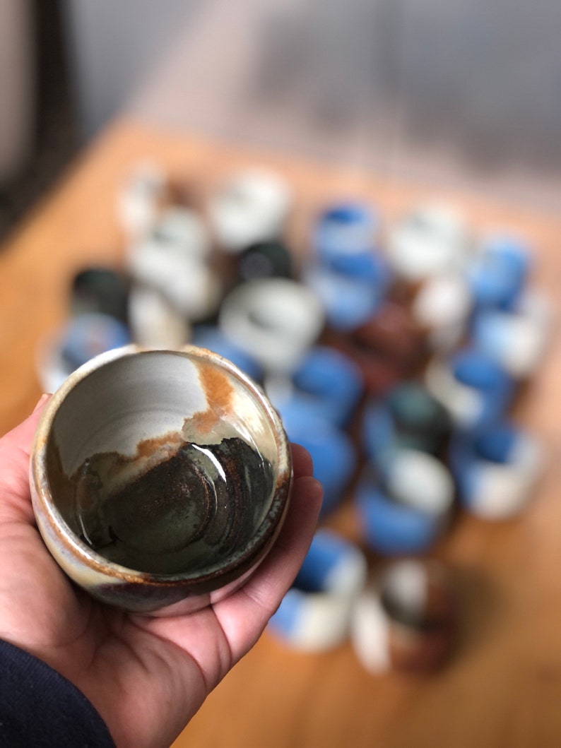 May include: A small, brown and white ceramic bowl with a dark brown glaze inside. The bowl is being held in a hand, and there are many other similar bowls in the background.