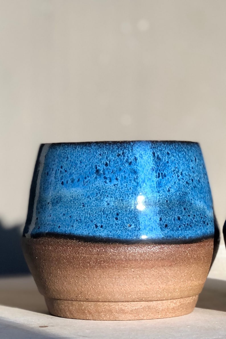 May include: A small, round ceramic mug with a blue glaze and a brown rim. The mug has a speckled texture and a glossy finish.