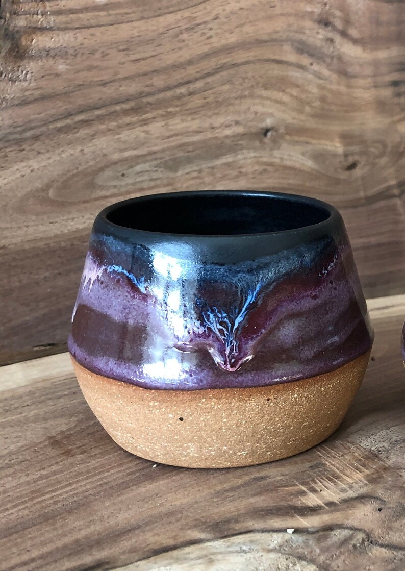 May include: A ceramic mug with a dark brown, purple, and blue glaze. The mug has a rounded shape and a wide base.