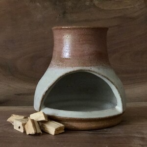 Puede incluir: Un quemador de incienso de cer&aacute;mica blanco y marr&oacute;n con una abertura redondeada. El quemador est&aacute; sobre una superficie de madera con varios trozos de madera de incienso a su lado.