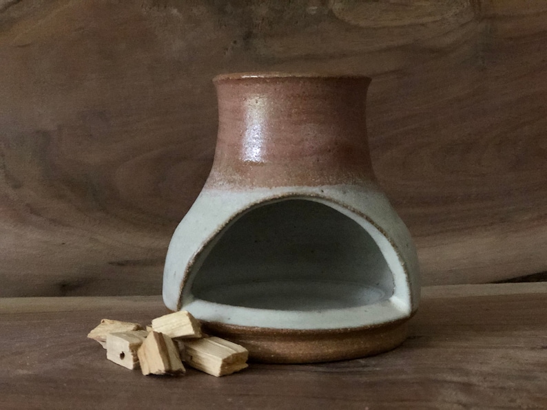 Puede incluir: Un quemador de incienso de cer&aacute;mica blanco y marr&oacute;n con una abertura redondeada. El quemador est&aacute; sobre una superficie de madera con varios trozos de madera de incienso a su lado.