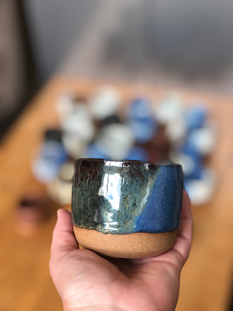 May include: A small, handmade ceramic cup with a blue, green, and brown glaze. The cup has a rounded shape and a slightly flared rim.