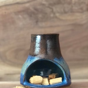 Puede incluir: Un quemador de incienso de cer&aacute;mica azul y marr&oacute;n con una peque&ntilde;a abertura en la parte superior. El quemador tiene una base redondeada y una abertura ancha en el centro. Hay varios peque&ntilde;os palitos de incienso de madera dentro del quemador.