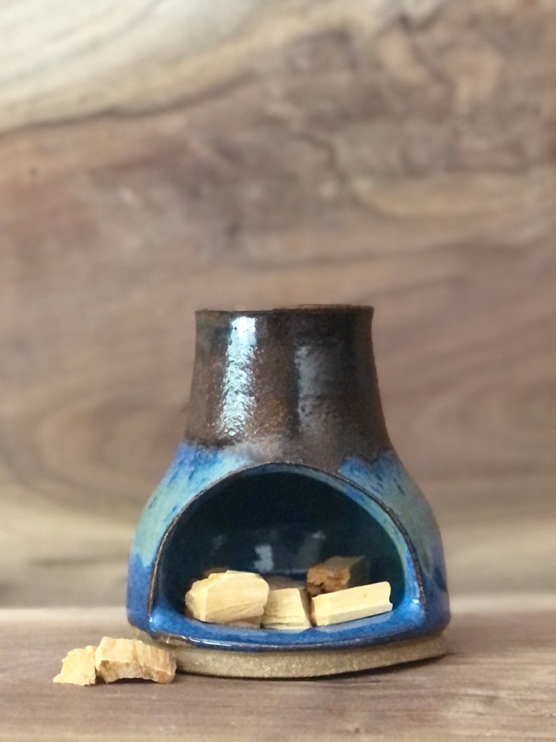 Puede incluir: Un quemador de incienso de cer&aacute;mica azul y marr&oacute;n con una peque&ntilde;a abertura en la parte superior. El quemador tiene una base redondeada y una abertura ancha en el centro. Hay varios peque&ntilde;os palitos de incienso de madera dentro del quemador.