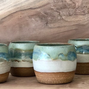 May include: Six ceramic mugs with a blue, green, and white glaze. The mugs are arranged in a row on a wooden surface.