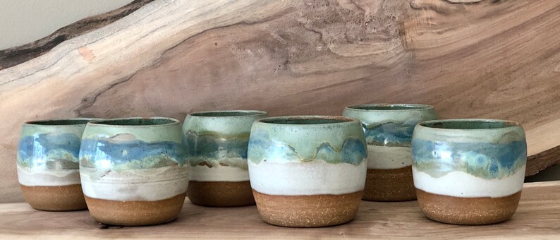 May include: Six ceramic mugs with a blue, green, and white glaze. The mugs are arranged in a row on a wooden surface.