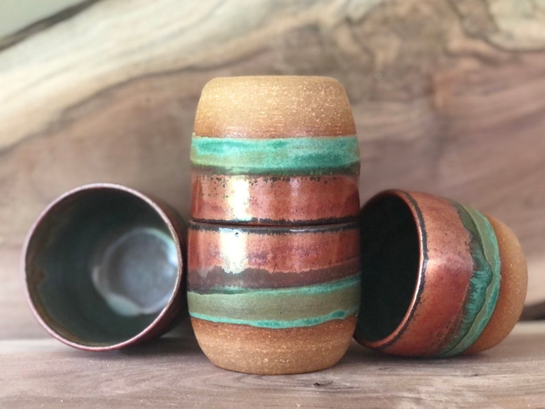 May include: Three ceramic mugs with a brown, copper, and green striped design. The mugs are stacked on top of each other.