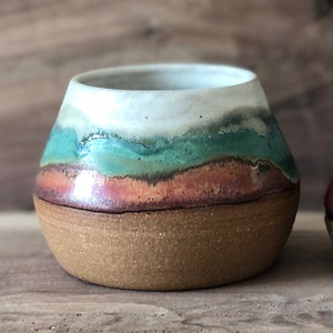 May include: A small, round ceramic pot with a white glaze and a layered design of green, orange, and brown. The pot has a rough, textured surface.