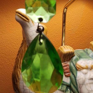May include: A green faceted crystal pendant hangs in front of a ceramic camel figurine. The pendant is a vibrant green color and has a teardrop shape. The camel is white with brown and green accents.