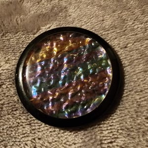 Könnte beinhalten: Ein rundes, irisierendes Glasobjekt mit schwarzem Rand. Das Glas hat eine strukturierte Oberfläche und reflektiert eine Vielzahl von Farben, darunter Gold, Grün, Lila und Blau. Das Objekt befindet sich vor einem neutralen Hintergrund.
